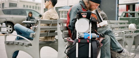 Links ein Mensch auf einer Bank mit Sonnenbrille, rechts ein offensichtlich Obdachloser mit seinen Habseligkeiten und Schlafsack