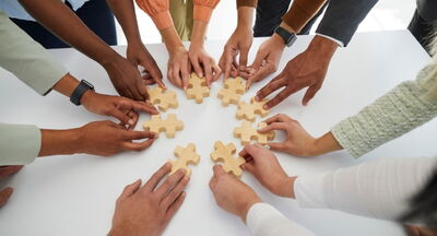 Viele Hände unterschiedlicher Hautfarbe kommen im Kreis zusammen, alle halten ein hölzernes Puzzleteil in der Hand