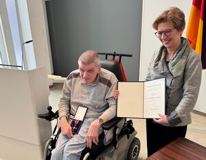 Ein älterer Mann im Rollstuhl, hält auf dem Schoß eine Schachtel mit einem Bundesverdienstkreuz, daneben eine Frau mit Urkunde lächelnd.