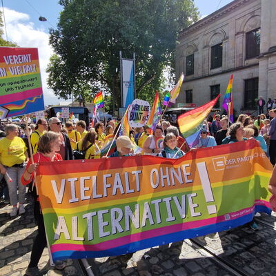 großes buntes Banner "Vielfalt ohne Alternative" und Logo des Paritätischen, dahinter viele Menschen mit bunten Flaggen bei einer Demo (CSD)