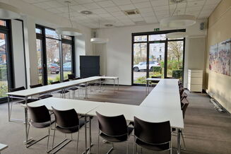 Heller Seminarraum mit weißen Tischen und ca. 15 Stühlen in Hufeisenform gestellt, Fenster zum Parkplatz, Bild an der Wand