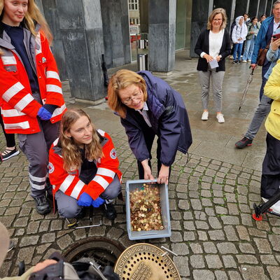 Mehrere Personen stehen um einen geöffneten Gullideckel, eine Frau hält eine Kiste mit Münzgeld in die Kamera