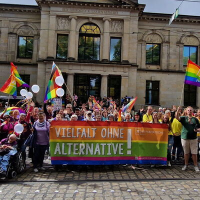 großes buntes Banner "Vielfalt ohne Alternative" und Logo des Paritätischen, dahinter und daneben viele Menschen mit bunten Flaggen bei einer Demo (CSD), stehend vor der Kunsthalle