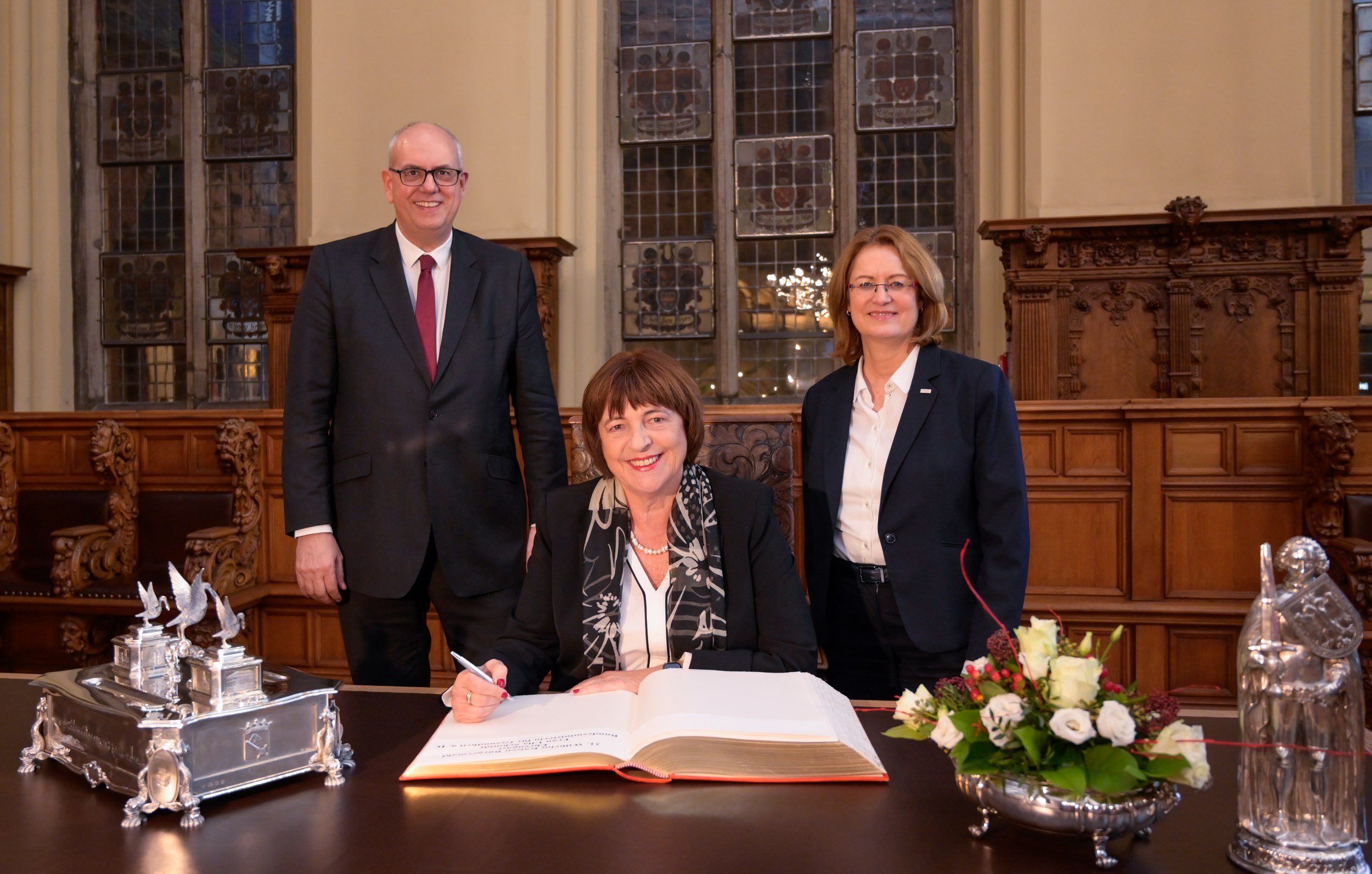 Bürgermeister und Bürgerschaftspräsidentin stehen hinter Ulla Schmidt, die mit einem Stift vor dem goldenen Buch sitzt